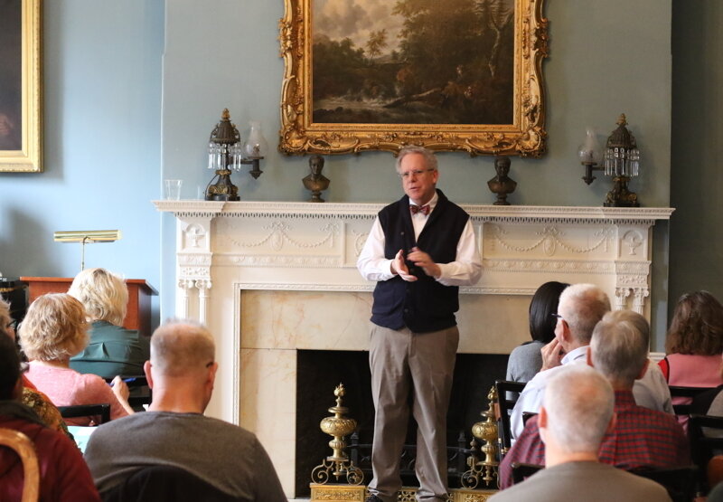 man giving a speech to a seated crowd in front of an ornate fireplace